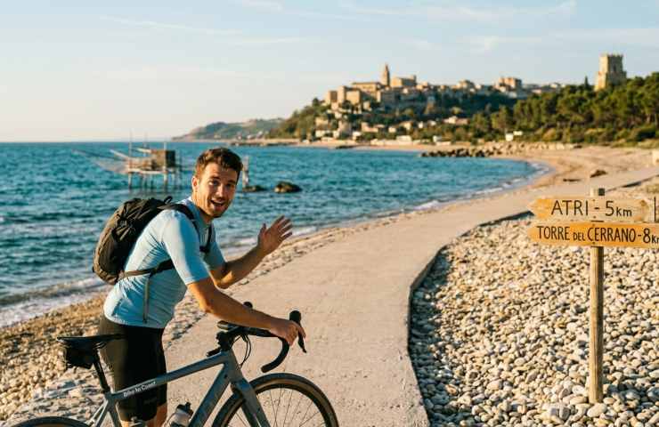 Ciclista in maglia azzurra saluta sulla pista ciclabile di Scerne di Pineto, Abruzzo, vicino a un cartello per Atri e la Torre del Cerrano sulla spiaggia di ciottoli.