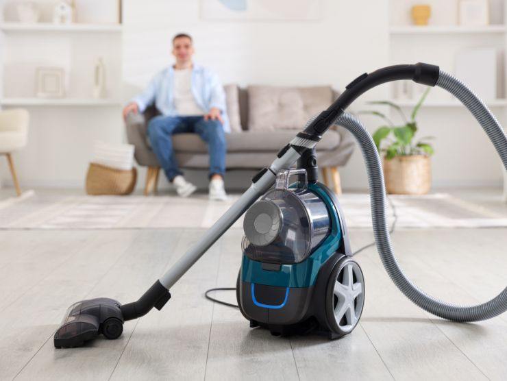 aspirapolvere in primo piano e uomo seduto sul divano come sfondo