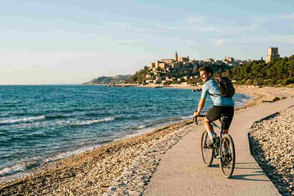 Cicloturista pedala sulla pista ciclabile Bike to Coast a Scerne di Pineto, Abruzzo, al tramonto, con vista sul borgo antico di Atri e la Torre del Cerrano.