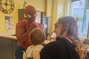 Uomo con costume di Spider-Man gioca con bimbo in braccio ad una donna sorridente.