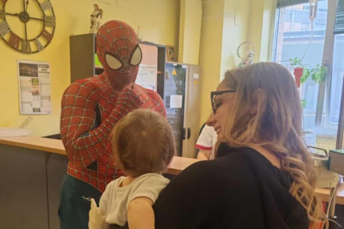 Uomo con costume di Spider-Man gioca con bimbo in braccio ad una donna sorridente.